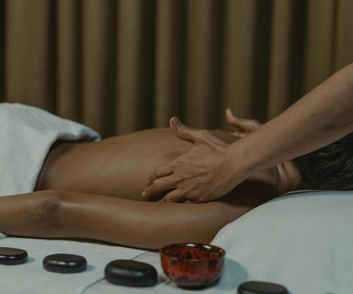 Vrouw ondergaat een ontspannende massage: de afbeelding toont de rug van de vrouw en de handen van de masseur tijdens de behandeling. Naast vrouw liggen hot stones die gebruikt worden tijdens de massage.